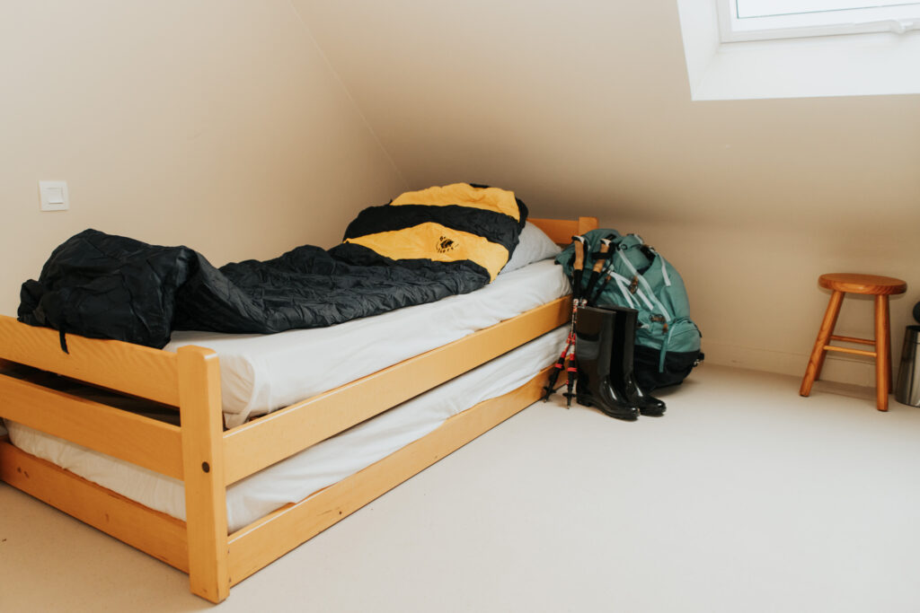 Un lit avec un sac de couchage. A côté il y a un sac de randonnée, des bottes et des bâtons de marche